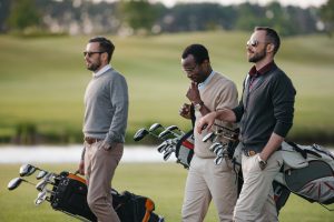 multiethnic golfers holding bags with golf clubs and walking on golf course